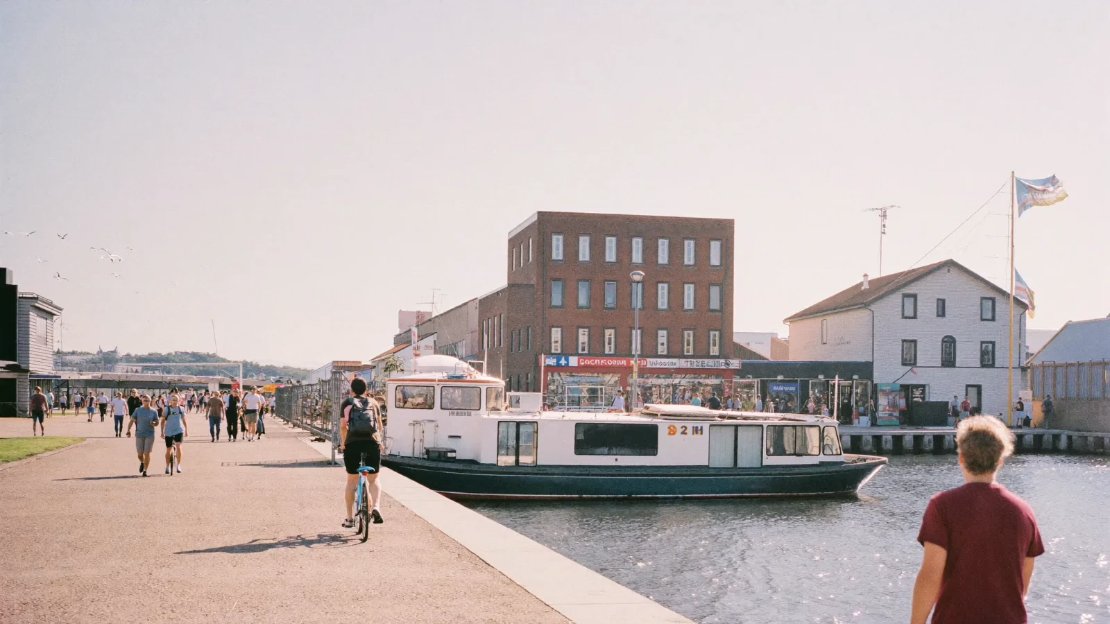 Hamnkaj i dagsljus där människor går och cyklar medan en passagerarfärja ligger vid bryggan