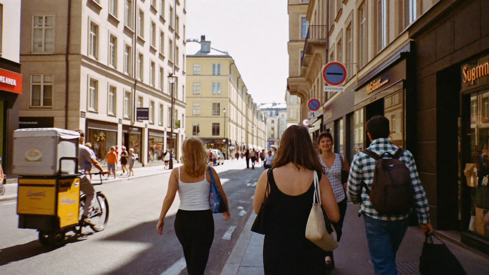 Livlig stadsgata mitt på dagen med människor som går förbi butiker i solljus och en cykelbud