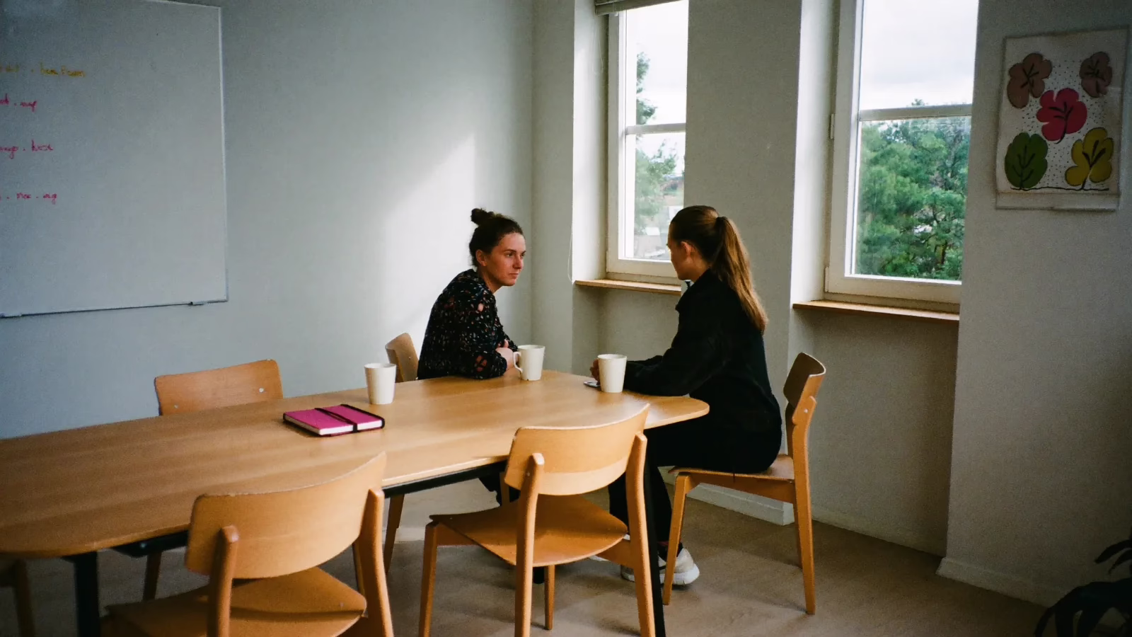 Två kollegor sitter i ett ljust mötesrum med kaffemuggar och pratar över bordet