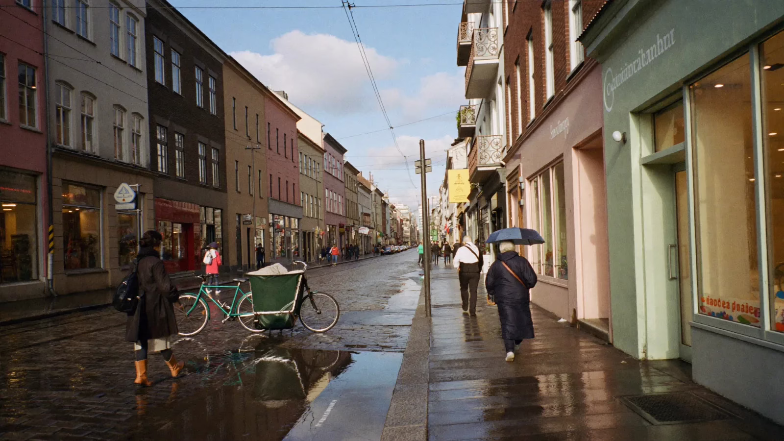 Nordisk gata efter regn där gatstenen speglar tända skyltfönster och människor går förbi