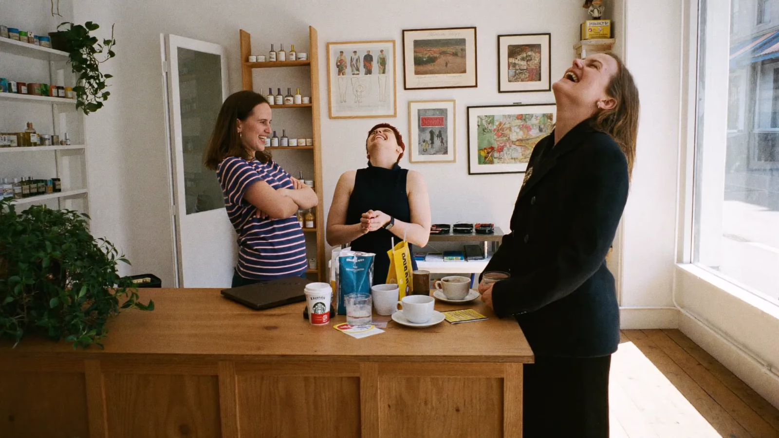 Tre kollegor står vid en butiksdisk och skrattar tillsammans med kaffemuggar på disken