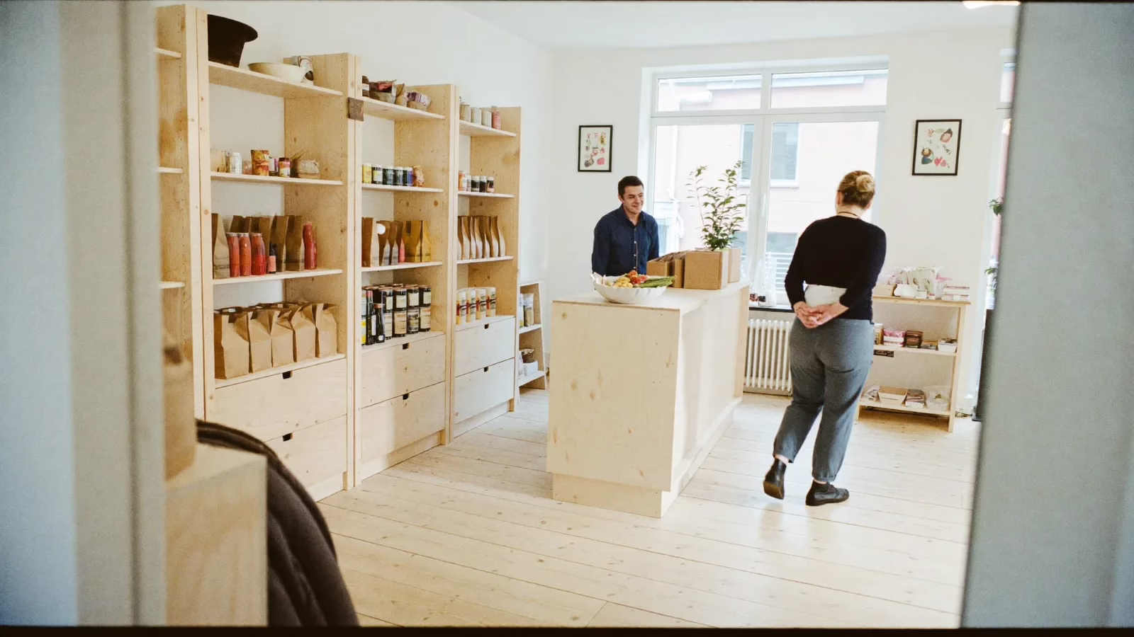 Ljus butik med björkhyllor där ägaren står vid disken och en kund kommer in genom dörren
