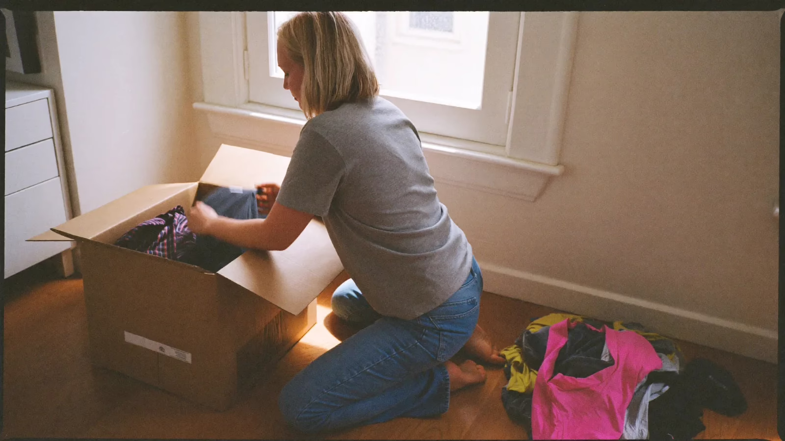 Person sitter på golvet och packar plagg i en kartong i ett ljust rum